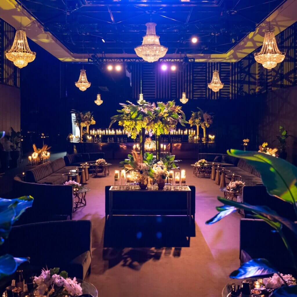 That Good Co The elegantly lit room, graced by chandeliers and a large floral centerpiece from That Good Co, features a table surrounded by candles and arranged seating.