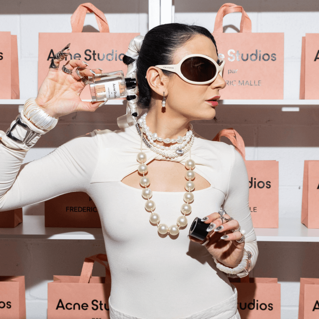 That Good Co Person wearing a white outfit, large sunglasses, and pearl accessories holds a That Good Co perfume bottle. Background features shelves with pink Acne Studios bags.