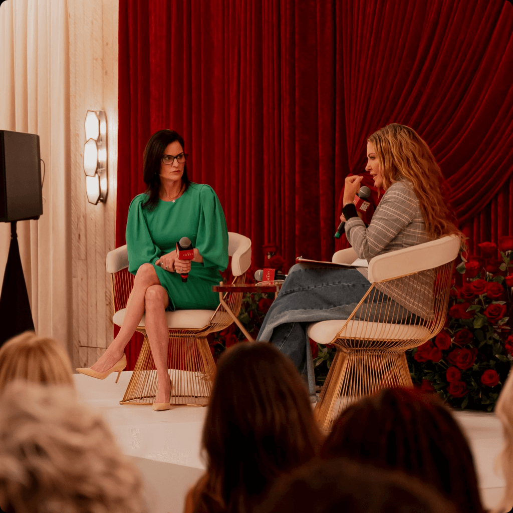 That Good Co Two women are sitting on stage, engaged in a conversation that seems to capture the spirit of That Good Co. One wears a green dress, the other a striped sweater and jeans. The backdrop is a rich red curtain, adding warmth to the scene as the audience watches in anticipation.