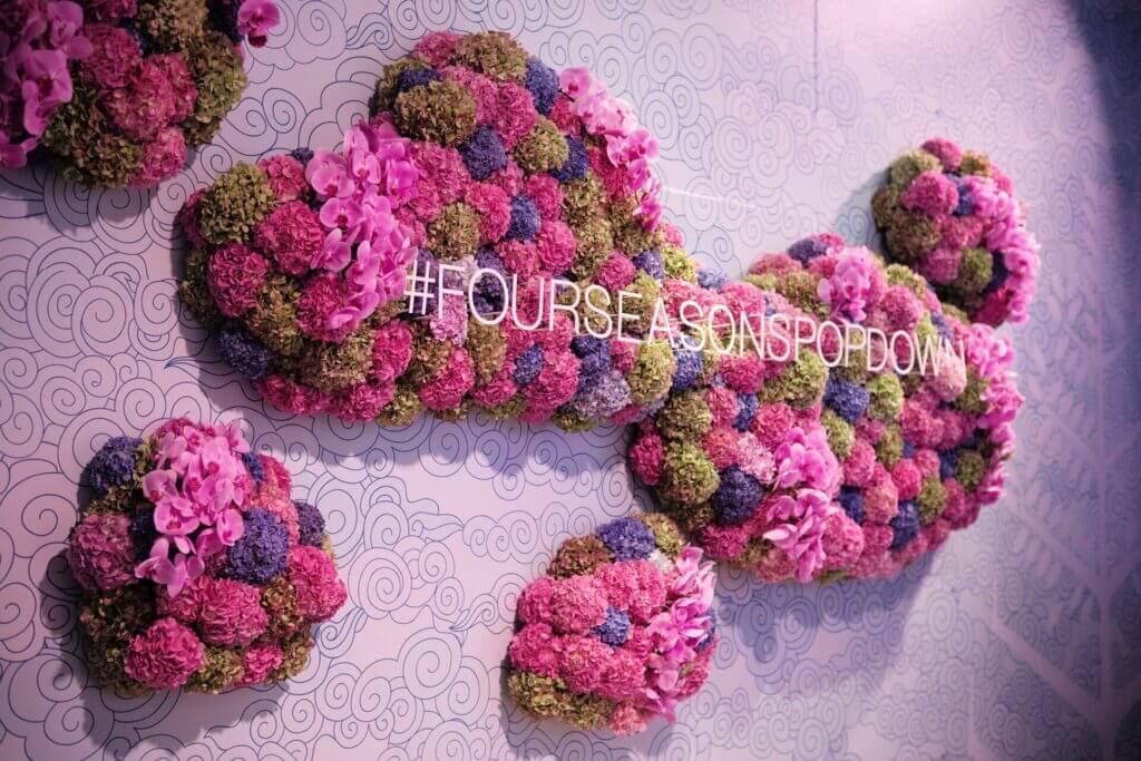 That Good Co A flower arrangement on a wall spells out "#FOURSEASONSPOPDOWN" with pink and green blooms on a patterned backdrop, inspired by That Good Co's artistic touch.