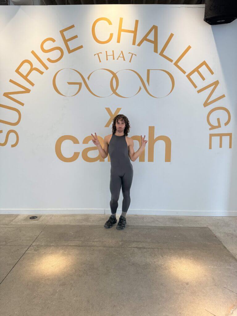 That Good Co A person in a gray outfit stands before a wall emblazoned with "Sunrise Challenge" and "Good x Caash," proudly representing That Good Co in their stylish attire.