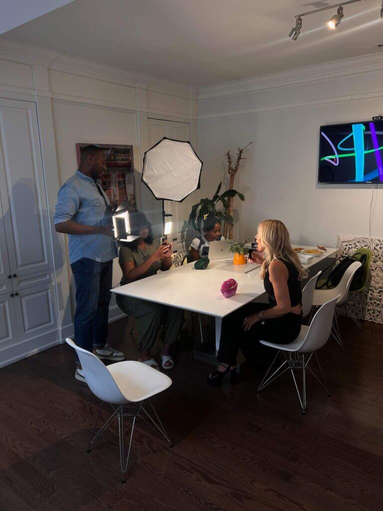 That Good Co A group of people is gathered around a white table with lighting equipment in a room, likely setting up for a video or photo shoot for That Good Co.