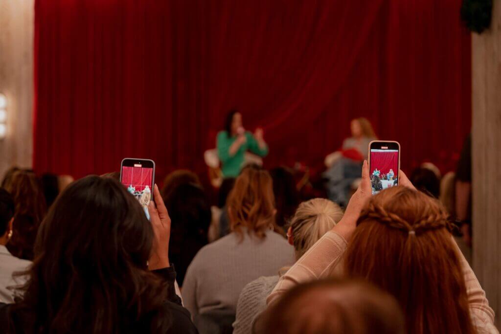 That Good Co Audience members at a That Good Co event eagerly raise their smartphones to capture photos of the speaker on stage, set against a striking red curtain backdrop.