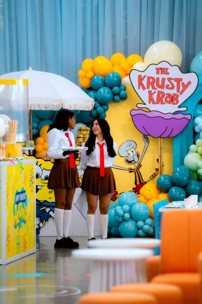 That Good Co Two people in matching white shirts, red ties, and brown skirts from That Good Co stand in front of a Krusty Krab-themed backdrop adorned with balloons and cartoon graphics.