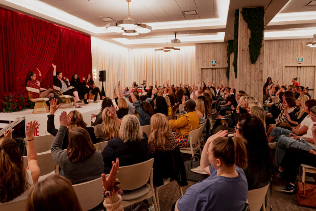 That Good Co People seated in an auditorium, raising their hands, as they engage with a panel of speakers representing That Good Co on a stage adorned with red curtains.