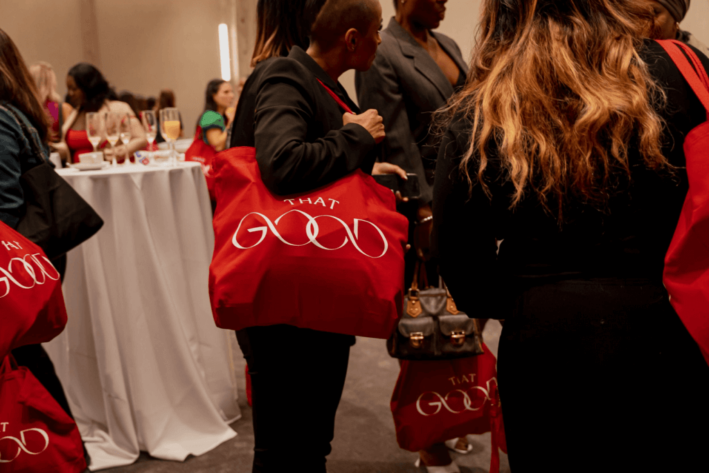 That Good Co People gathered at an event holding red tote bags emblazoned with "THAT GOOD" from That Good Co. Glasses of drinks are seen on a round table in the background.