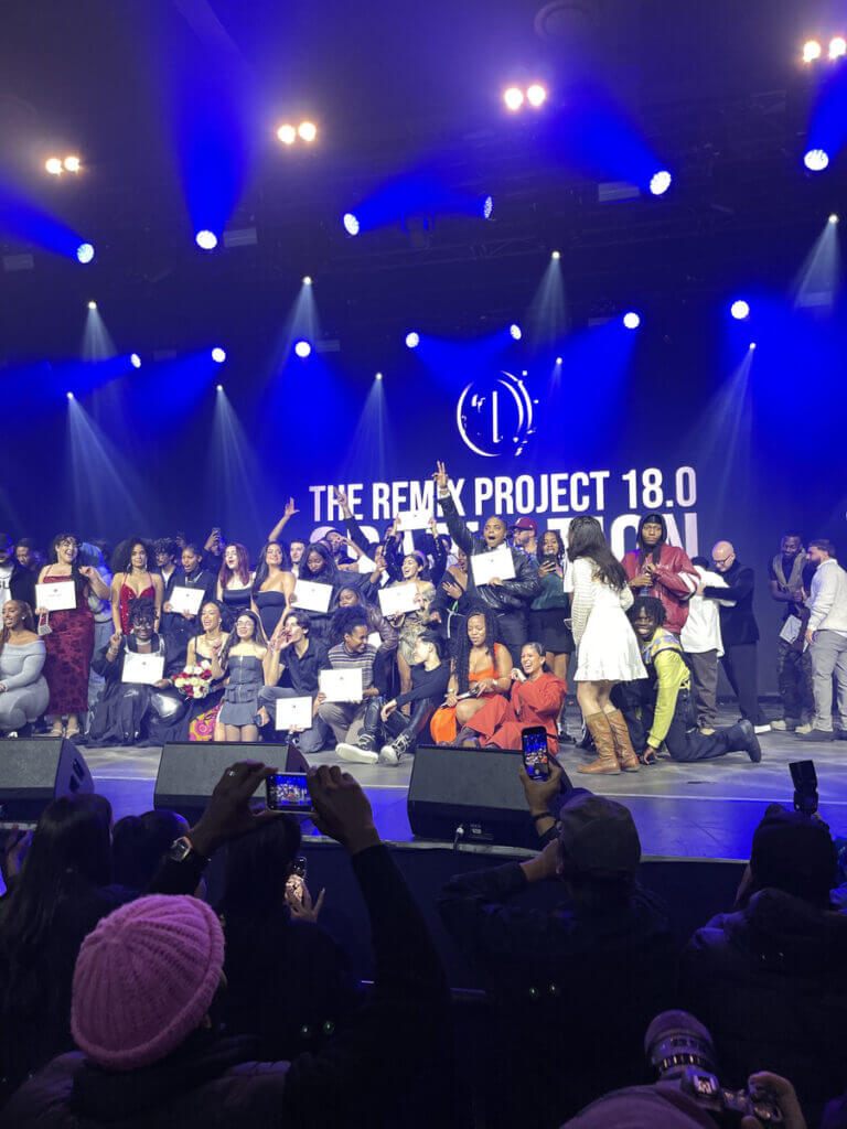 That Good Co A large group of people from That Good Co pose on stage under bright lights, holding certificates, with a "The Remix Project 18.0" banner in the background. Audience members eagerly snap photos, capturing the celebratory moment.