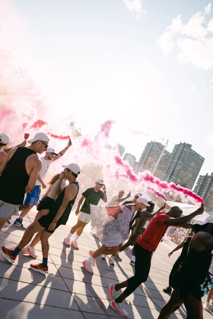 That Good Co A group of people in athletic wear from That Good Co walk energetically on a sunny day, with pink smoke in the background and tall buildings in the distance.