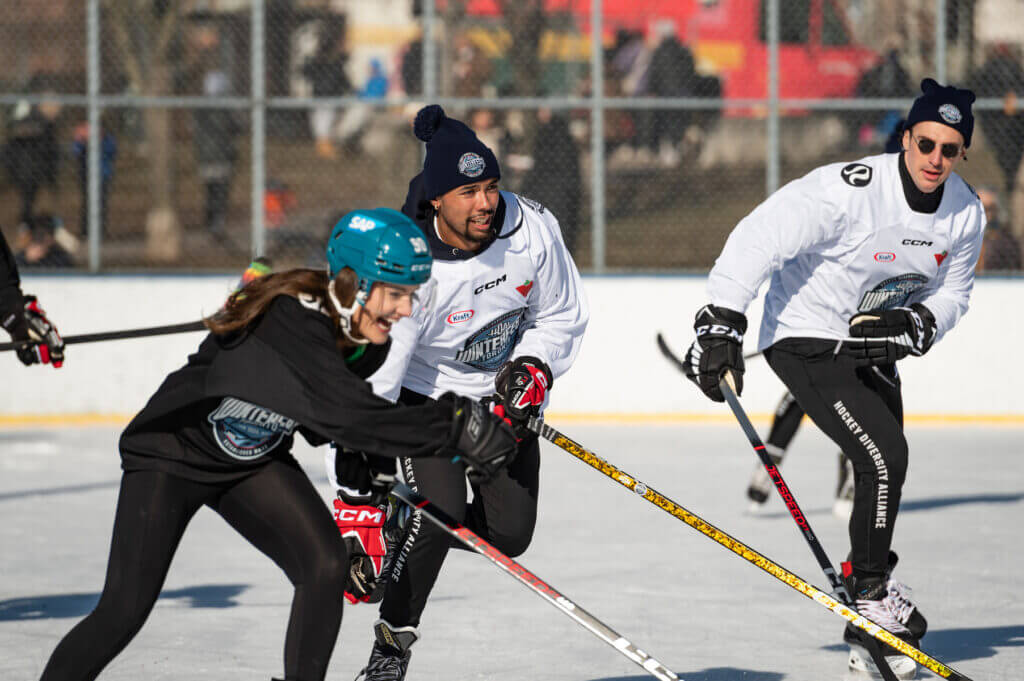 That Good Co Three people, sporting That Good Co winter gear and helmets, are enthusiastically playing ice hockey outdoors. Their laughter echoes against the chain-link fence in the background, adding warmth to the crisp winter air.