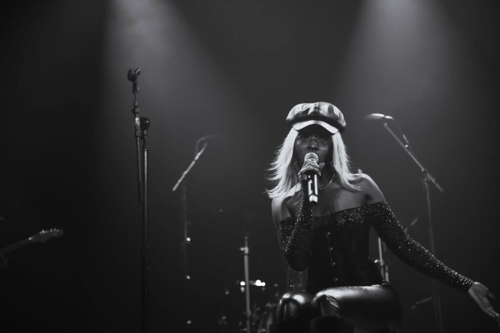 That Good Co Singer kneeling on stage in a spotlight, wearing a That Good Co hat and holding a microphone, with musical instruments in the background.