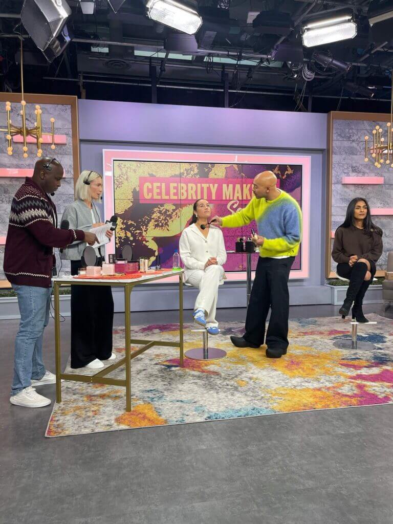 That Good Co Makeup artists from That Good Co are expertly applying makeup to a seated woman on a brightly lit studio set, where the glowing words 'Celebrity Makeover' shimmer across a screen in the background.