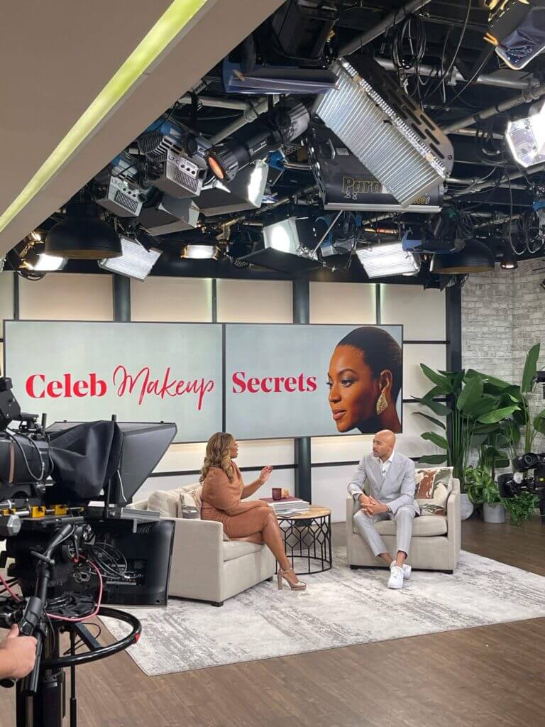 That Good Co A talk show set with two hosts from That Good Co. sitting on a couch, divulging makeup secrets. Cameras and studio lights illuminate the scene, while a large screen in the background proudly displays the show's title.