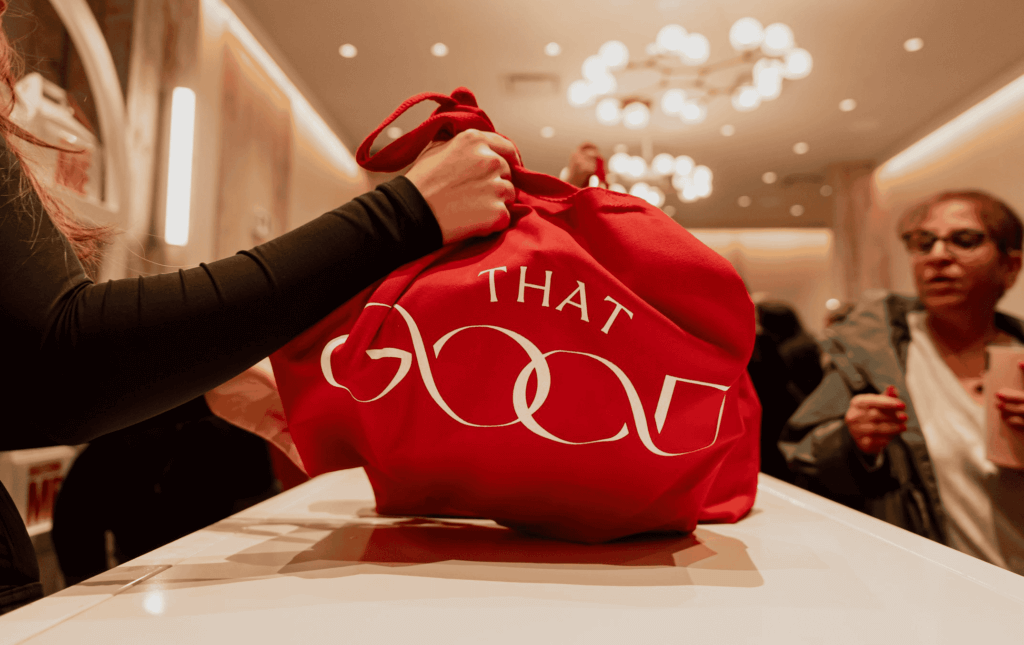 That Good Co A person holds a red bag emblazoned with "THAT GOOD" in a warmly lit room bustling with others, reflecting the signature style of That Good Co.