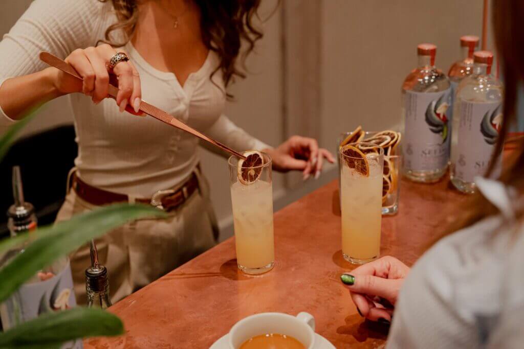 That Good Co A patron at the bar expertly places a dried citrus slice into their cocktail glass, proudly sipping That Good Co's finest creation. Bottles and a cup scatter the counter, each adding to the vibrant ambiance of craftsmanship.