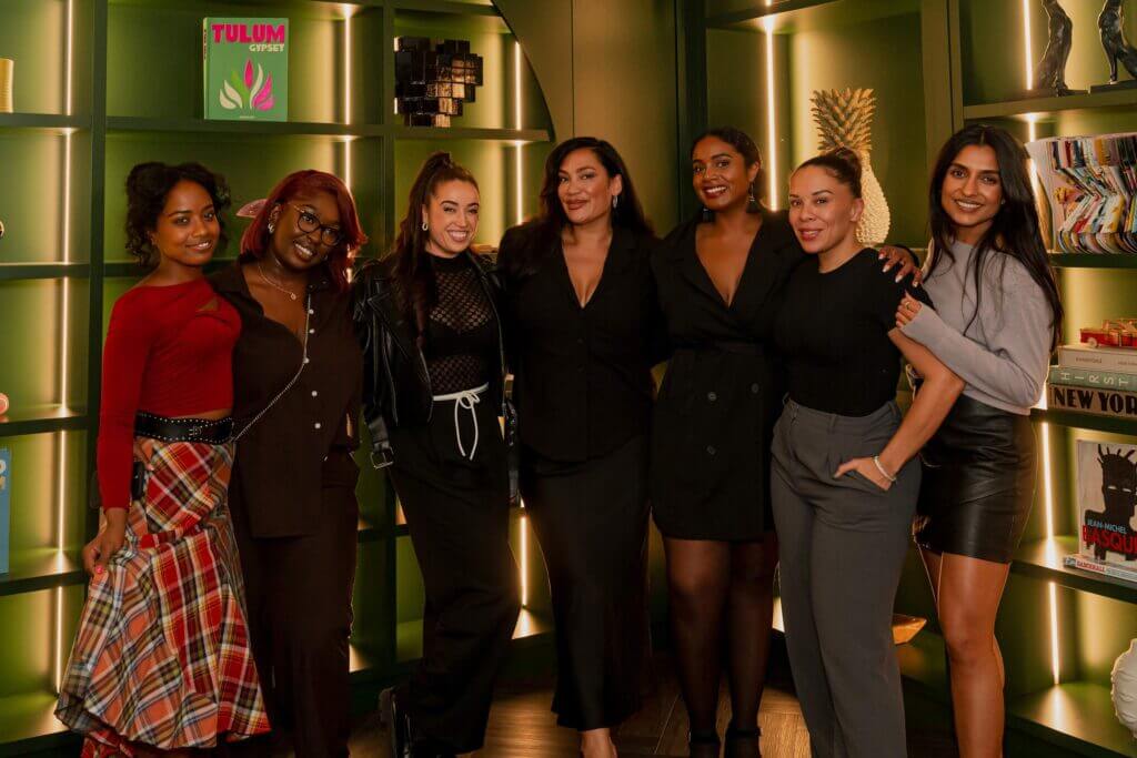 That Good Co Seven women from That Good Co smile in front of a bookshelf adorned with lights, each showcasing unique and stylish outfits.