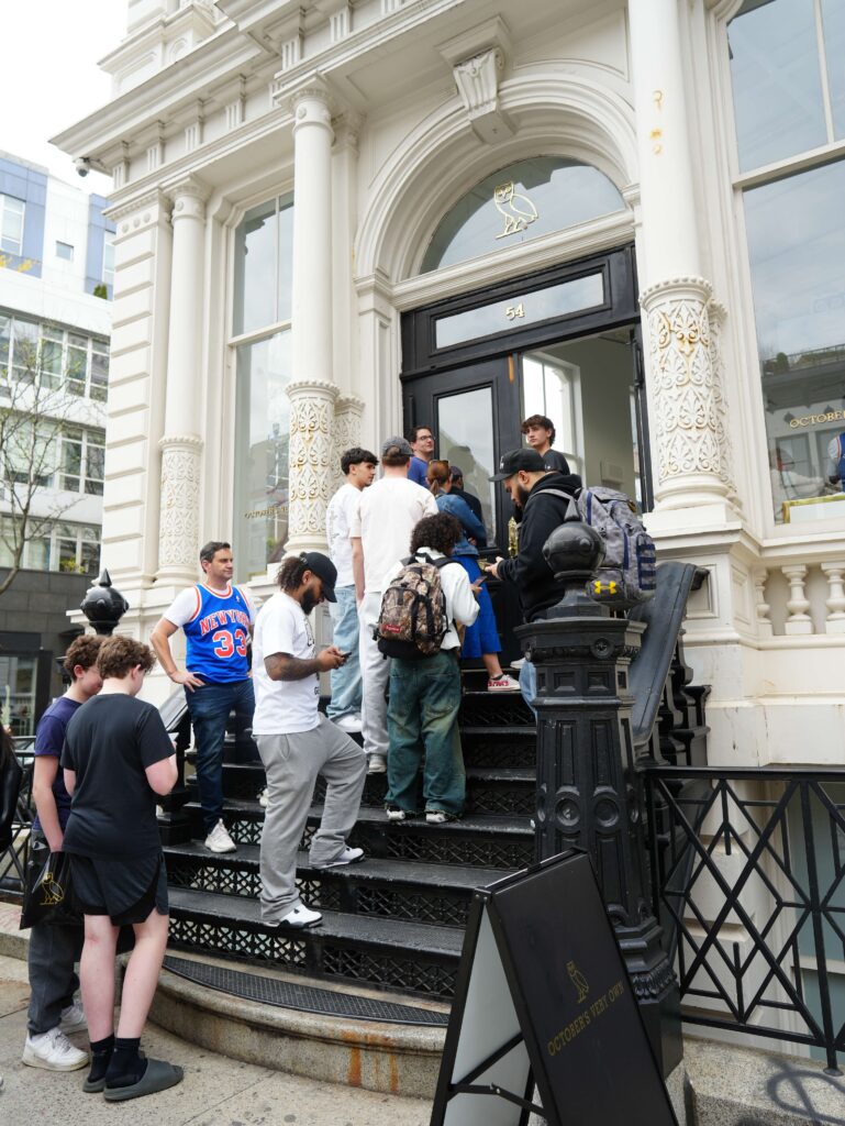 That Good Co A group of people wait in line on the steps outside a storefront with ornate white columns and a large arched doorway.