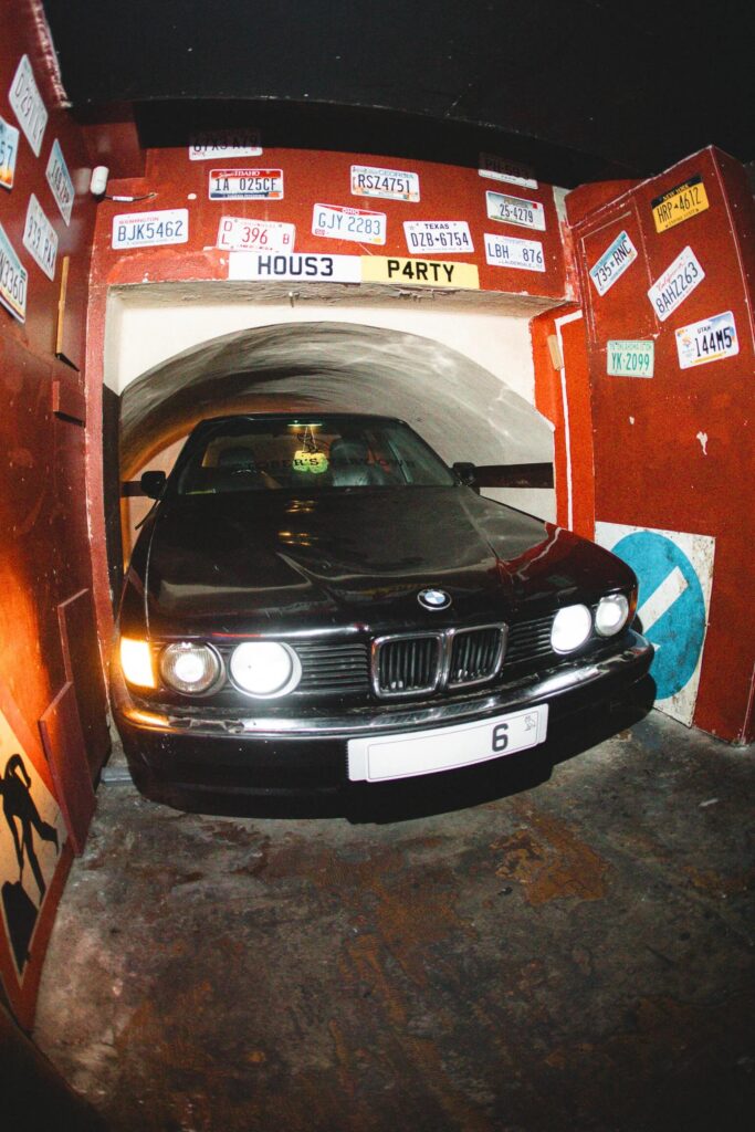That Good Co A black BMW car parked inside a tunnel-like garage decorated with various license plates and street signs on red walls under a sign that reads "House Party.
