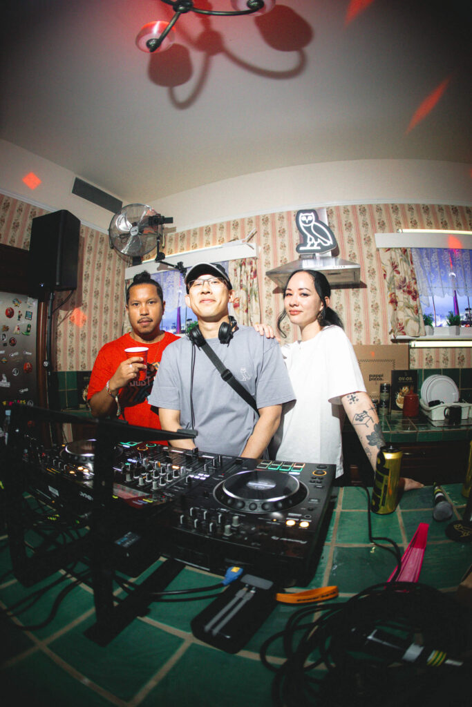 That Good Co Three people stand behind a DJ setup in a kitchen with patterned wallpaper, dishes on the counter, and a neon sign on the wall.
