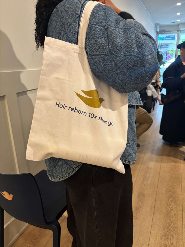 That Good Co Person carrying a white tote bag with a yellow dove logo and the text "Hair reborn 10x Stronger" while standing indoors near a group of people.