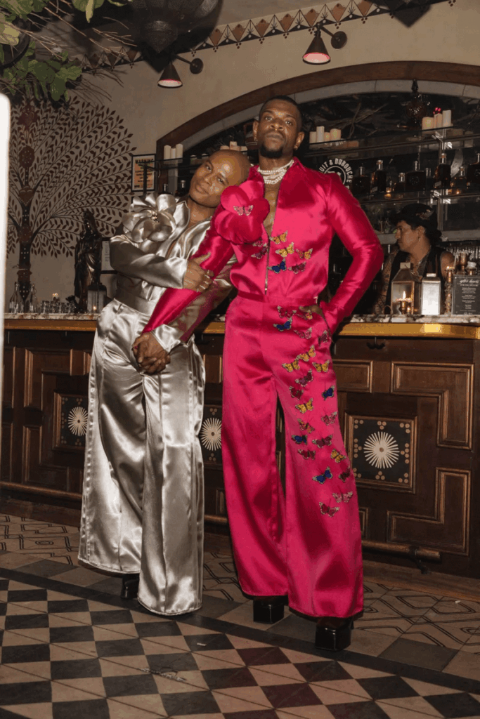 That Good Co Two people stand together in a bar, wearing shiny, wide-legged jumpsuits—one silver, one fuchsia with butterfly embellishments—on a patterned tile floor.