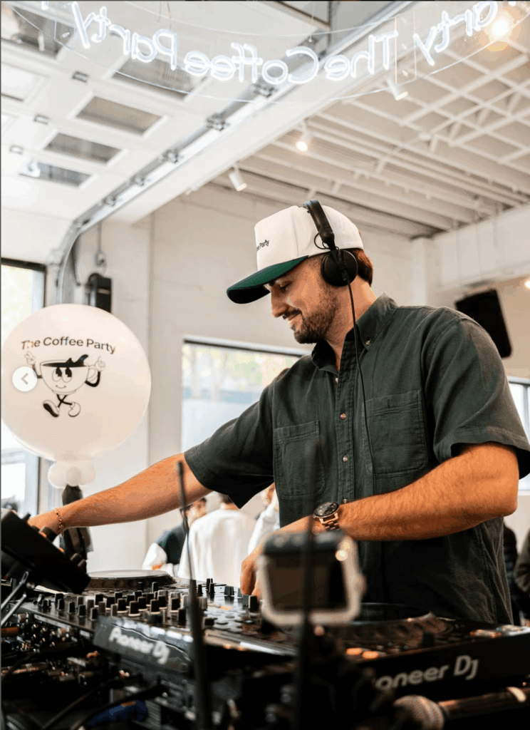 That Good Co A man wearing headphones and a baseball cap operates DJ equipment at an indoor event with a "The Coffee Party" balloon in the background.