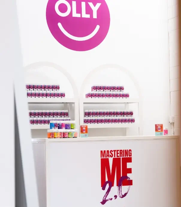 That Good Co A display area with shelves of OLLY supplement containers and a counter labeled "MASTERING ME 2.0" in pink and red text. The OLLY logo is visible on the wall above.