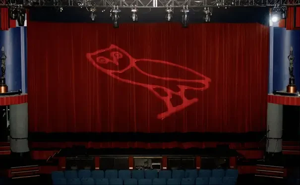 That Good Co A red theater curtain with a simple owl logo projected onto it. The stage is empty and seats are visible in the foreground.