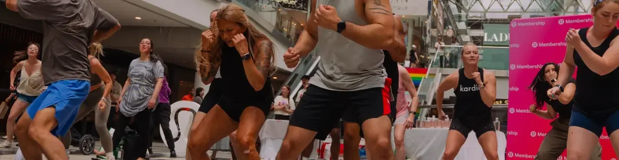 That Good Co A group of people participate in a fitness or dance class inside a shopping mall, with some spectators and a Membership sign in the background.