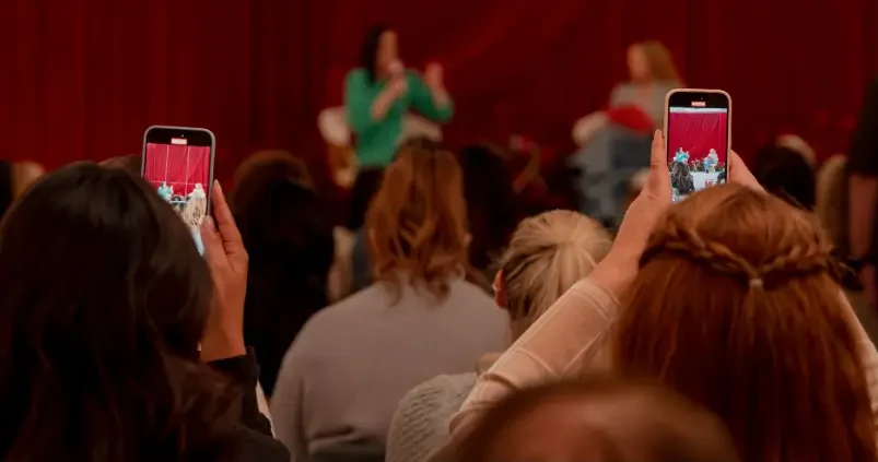That Good Co Audience members hold up phones to record two women speaking on a stage with a red curtain backdrop.