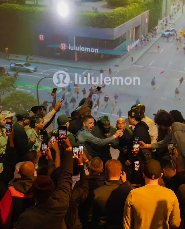 That Good Co A crowd of people take photos and videos with their phones in front of a large screen displaying a Lululemon store and the Lululemon logo.