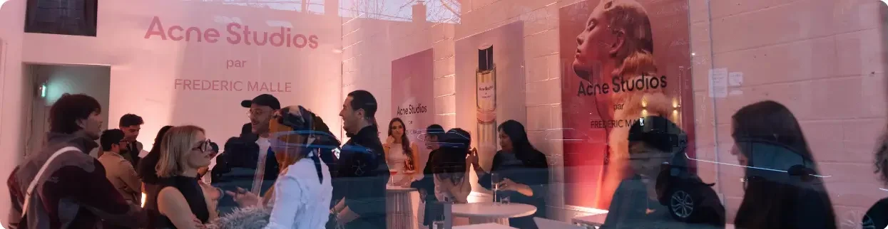 That Good Co People gather inside a store with pink lighting, featuring large posters for Acne Studios and Frederic Malle on the walls.