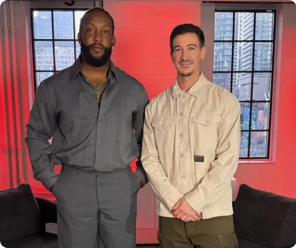 That Good Co Two men stand side by side indoors in front of large windows, with a cityscape visible in the background and red lighting in the room.