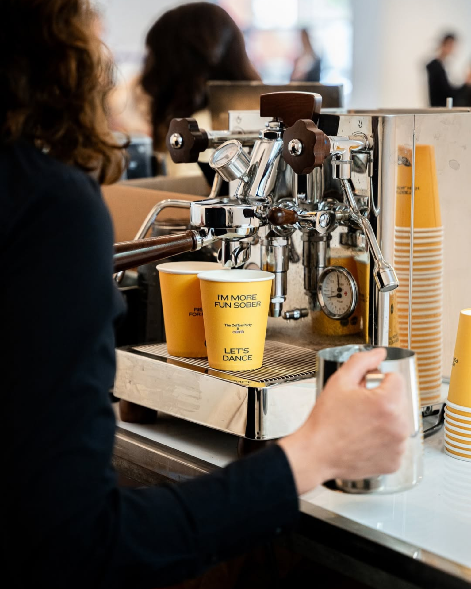 That Good Co Person operating an espresso machine, preparing a coffee drink in a yellow cup that reads "I'M MORE FUN SOBER" and "LET'S DANCE"; stacks of similar cups are nearby.